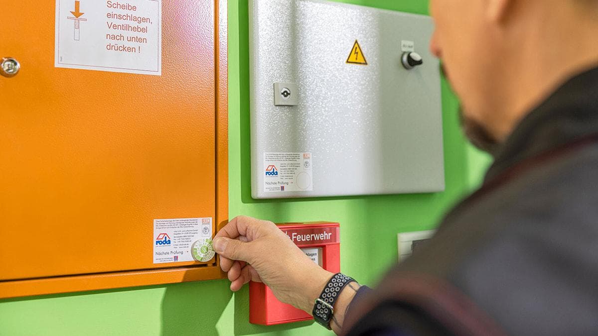Hand an einer Brandschutzeinrichtung, daneben zwei Prüfplaketten mit dem Vermerk "Nächste Prüfung".